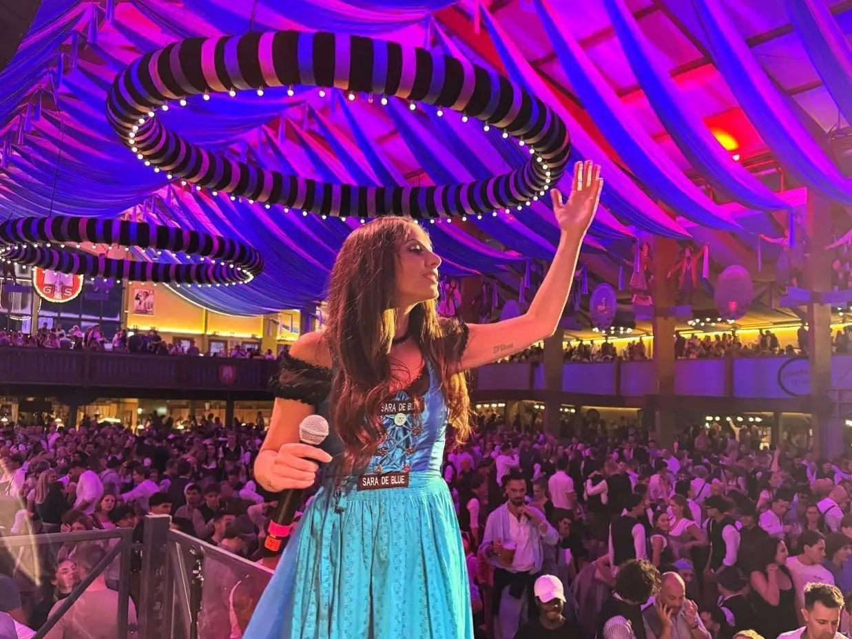 Eine Frau in einem blauen Dirndl steht mit einem Mikrofon auf einer Bühne und winkt einer großen Menschenmenge in einem festlich beleuchteten Festzelt zu.