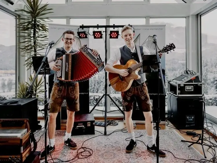 Zwei junge Musiker in traditioneller Tracht spielen in einem lichtdurchfluteten Raum mit Alpenpanorama im Hintergrund. Einer spielt Akkordeon, der andere Gitarre, beide stehen lächelnd auf einem Teppich vor Mikrofonen und Lautsprechern.